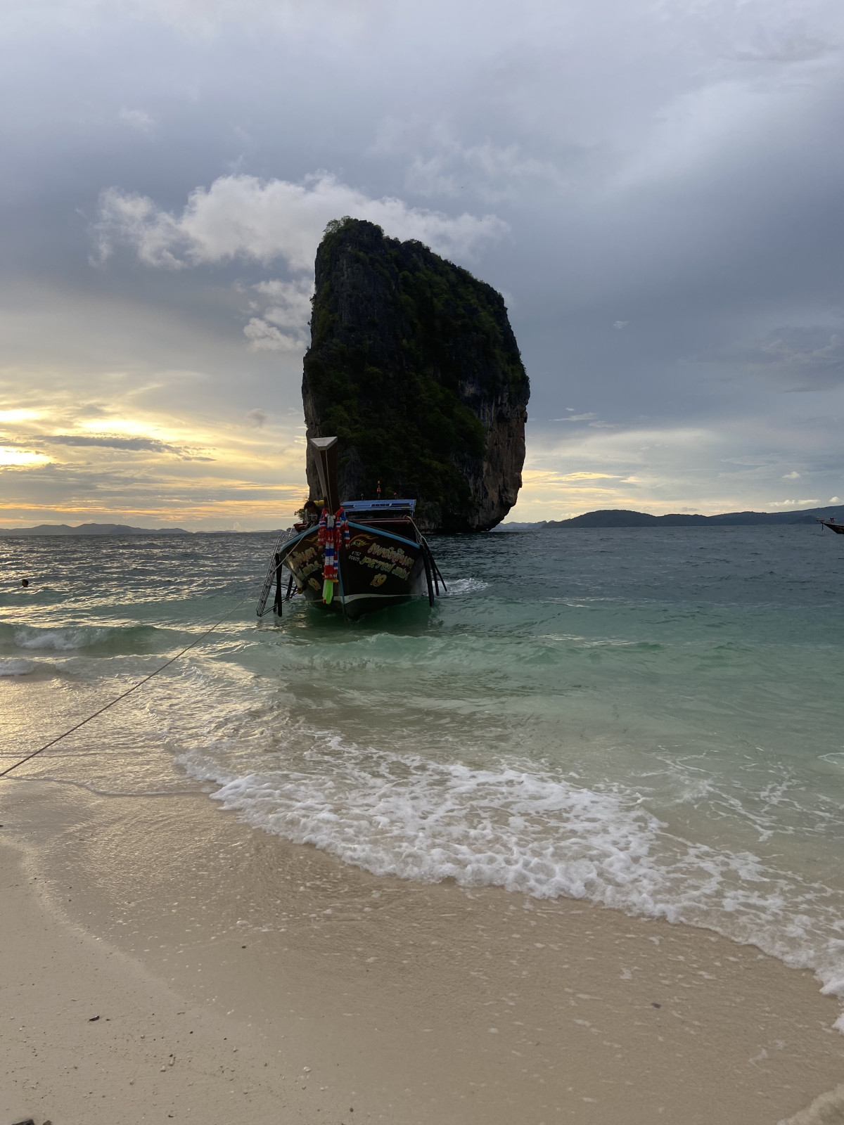 A long-tail boat rests on the shore of a beautiful beach in Thailand as the sun sets behind a towering limestone cliff