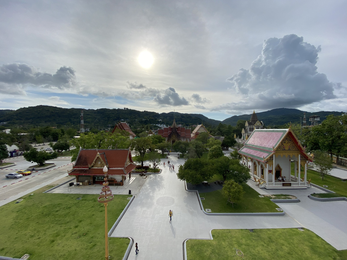 An elevated view of a tranquil temple complex in Thailand, featuring traditional architecture and lush green grounds under a bright, hazy sun