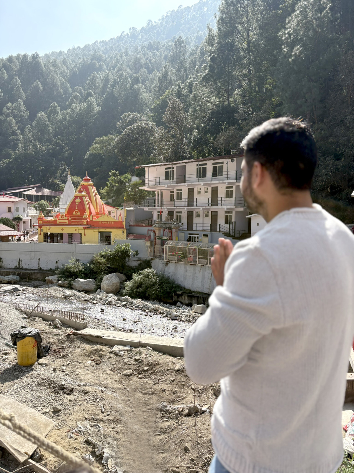 A man in a white sweater stands with his hands clasped in a gesture of prayer, looking out at a vibrant red and yellow Hindu temple