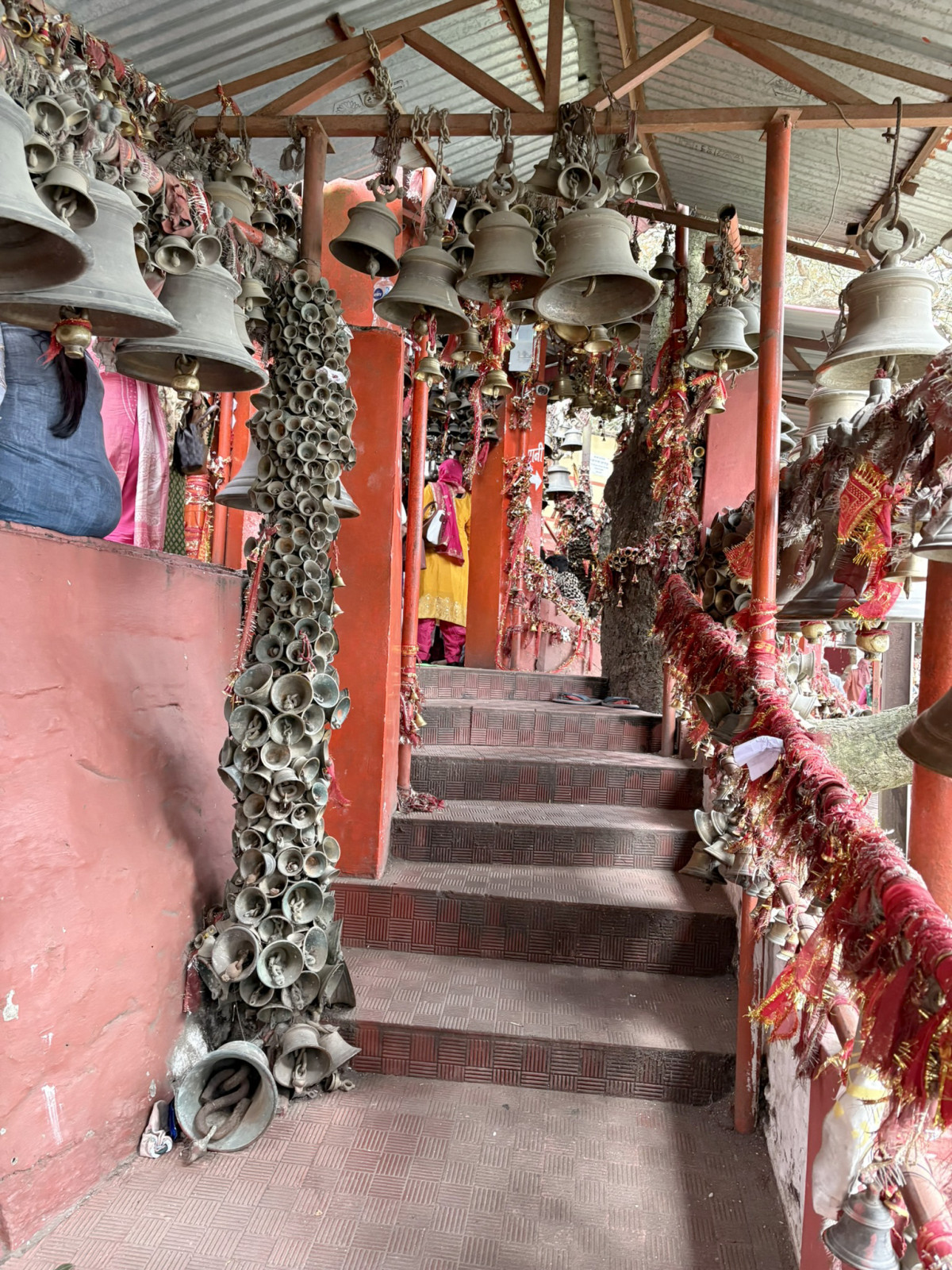 A flight of red-toned steps leading up within a Hindu temple that is heavily decorated with hundreds of brass bells and red ritual threads hanging from the ceiling and wrapped around the railings and pillars