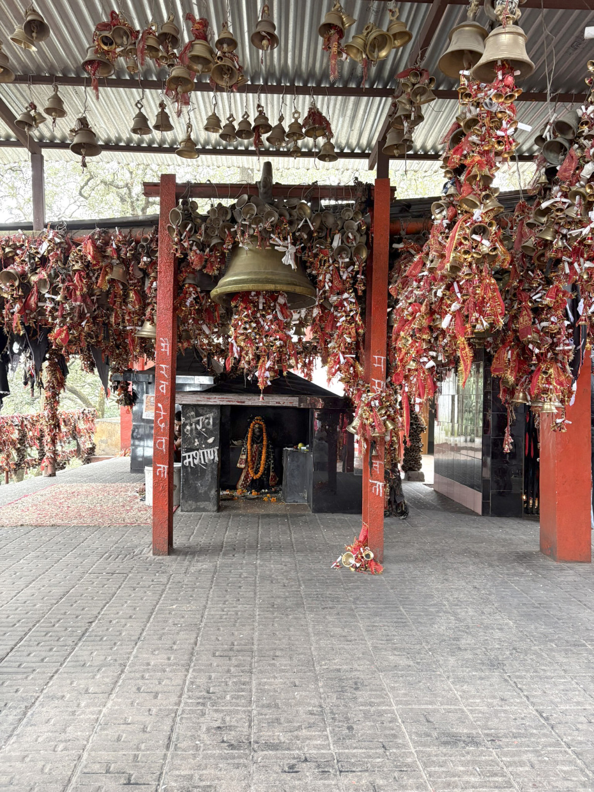 The courtyard of a Hindu temple densely decorated with hundreds of small brass bells and red and yellow ritual threads, hanging from the corrugated metal roof and pillars, with a small inner shrine visible in the background