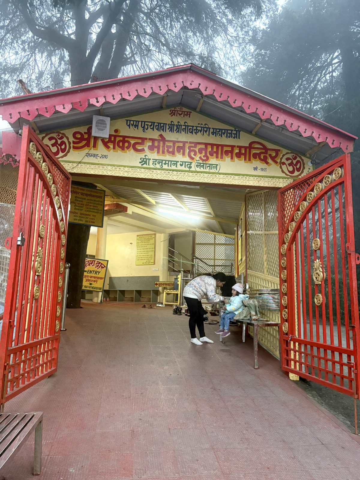 The brightly painted red and yellow ornate gateway and entrance to the Shri Sankat Mochan Hanuman Mandir on a foggy or misty day, with a woman attending to a child near the entrance bench
