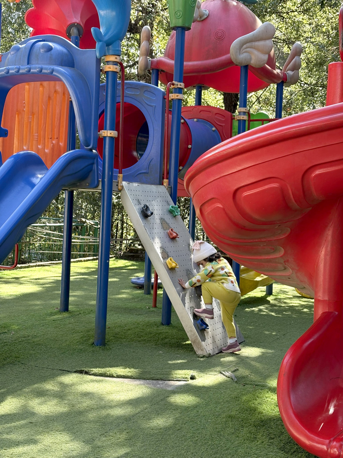 A small child in a yellow outfit and sun hat is actively climbing a miniature rock wall feature on a brightly colored, large outdoor playground with slides and a soft green ground surface