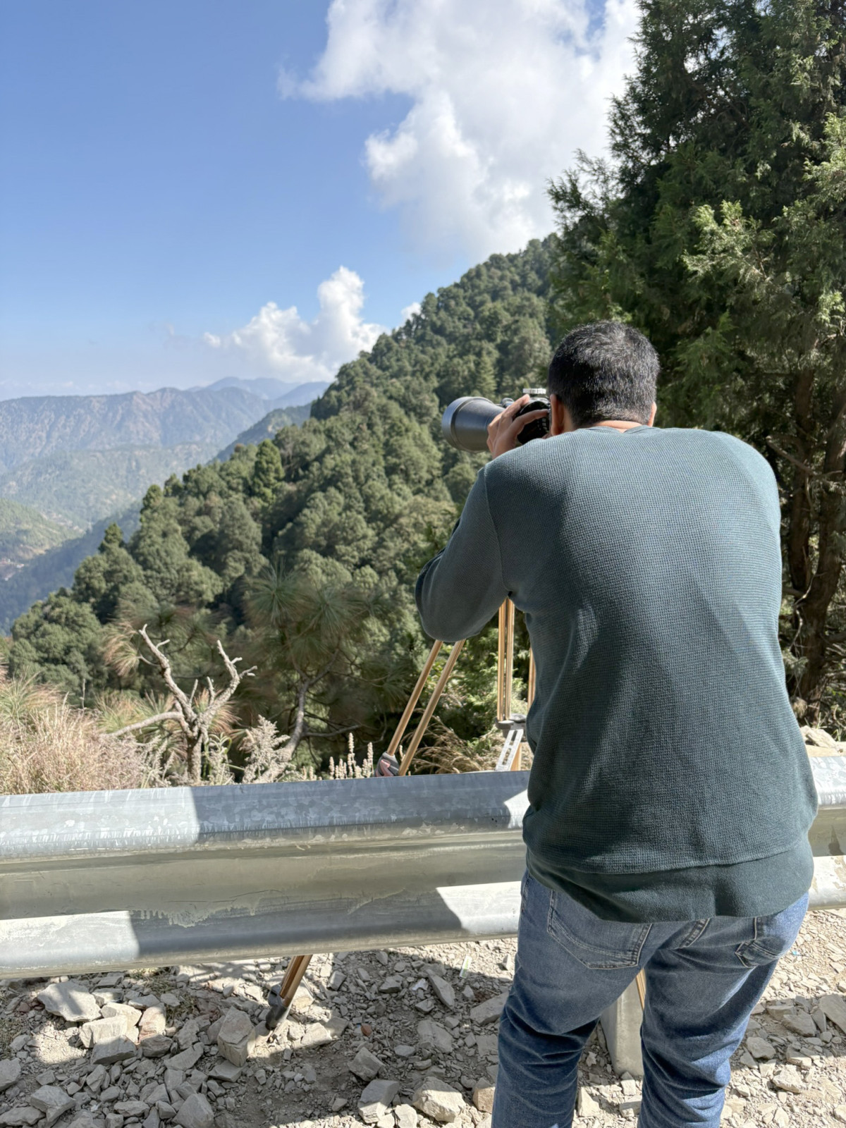 A man in a dark green sweater and jeans is looking through a viewfinder or telescope mounted on a tripod, observing the mountain range covered in dense green forest below a bright, sunny sky